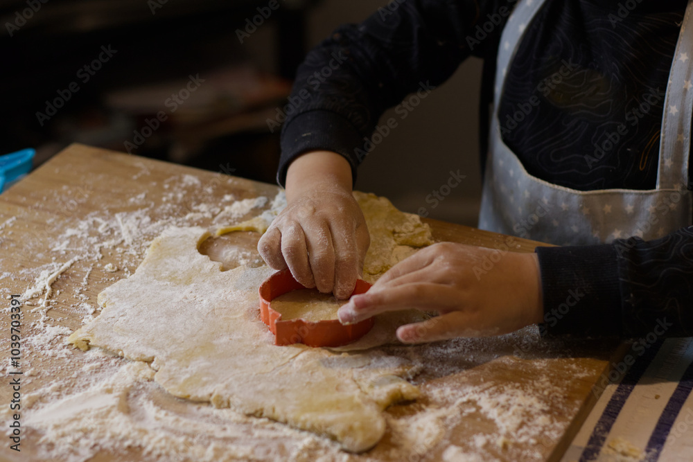 Kid making fancy Halloween biscuits with cookie dough dinosaur pumpkin cutter on a messy floured wooden board. Kid hands closeup on black shirt and apron. Kid making fancy Halloween biscuits with cookie dough dinosaur pumpkin cutter on a messy floured wooden board. Kid hands closeup on black shirt and apron.