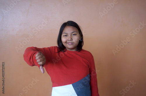 Dissatisfied Asian Little Girl Showing Thumb Down Dislike Gesture Isolated On Brown Background  photo