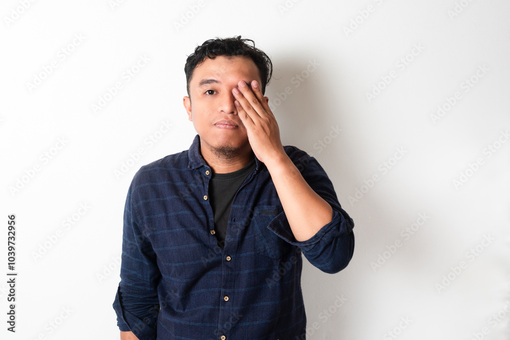 Asian man covers one eye with his hand, expressing fatigue or frustration. His neutral, yet tired expression suggests stress or contemplation, making this image ideal for themes of emotion, burnout Asian man covers one eye with his hand, expressing fatigue or frustration. His neutral, yet tired expression suggests stress or contemplation, making this image ideal for themes of emotion, burnout