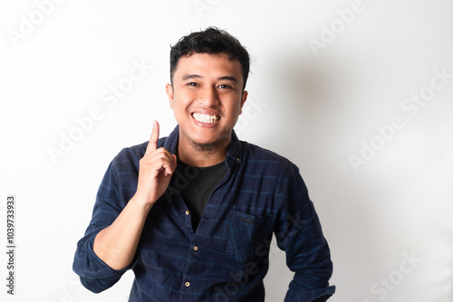 Adult Asian man looking at camera with happy expression while pointing up with one finger photo