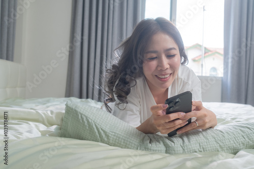 A woman lying comfortably on a bed, smiling while focused on her smartphone and tablet. The bright and cozy atmosphere captures a cheerful moment of happiness in her personal space.