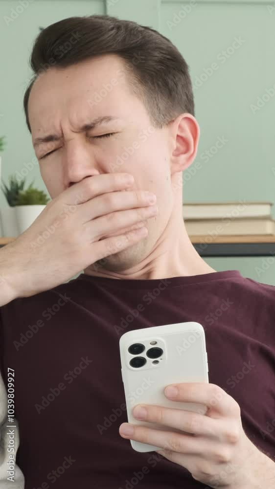Bored Caucasian young adult man using smartphone and yawning while spending leisure time at home. Concept of a boredom and tiredness. Vertical video. Bored Caucasian young adult man using smartphone and yawning while spending leisure time at home. Concept of a boredom and tiredness. Vertical video.