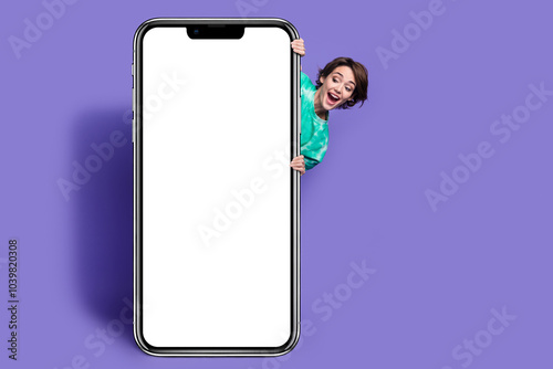 Full size photo of young woman behind big smartphone look empty space dressed stylish green clothes isolated on violet color background