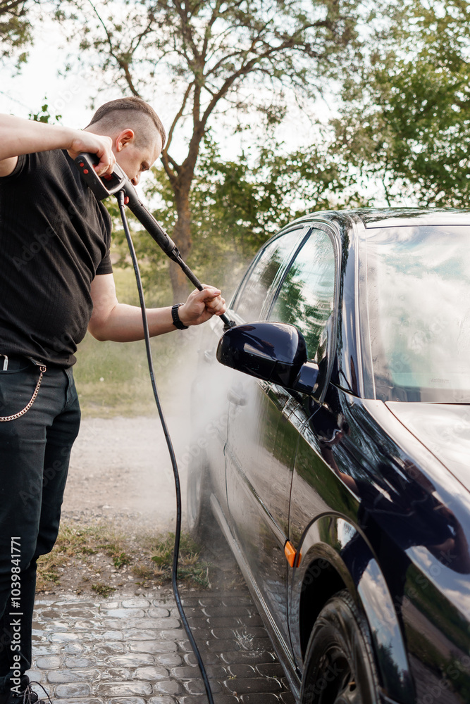 Man washing a car with a high-pressure washer on a sunny day in a green outdoor setting Man washing a car with a high-pressure washer on a sunny day in a green outdoor setting