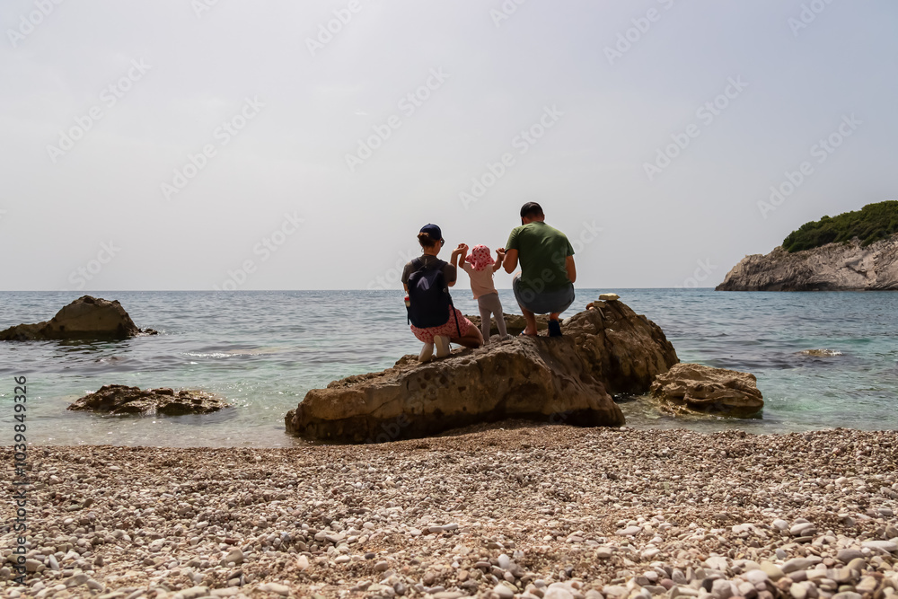 Caring parents with toddler on rock along idyllic pebble beach Filikuri in Himare, Albania. Crystal-clear turquoise water of Ionian Mediterranean sea. Concept family vacation in summer. Happy emotion Caring parents with toddler on rock along idyllic pebble beach Filikuri in Himare, Albania. Crystal-clear turquoise water of Ionian Mediterranean sea. Concept family vacation in summer. Happy emotion