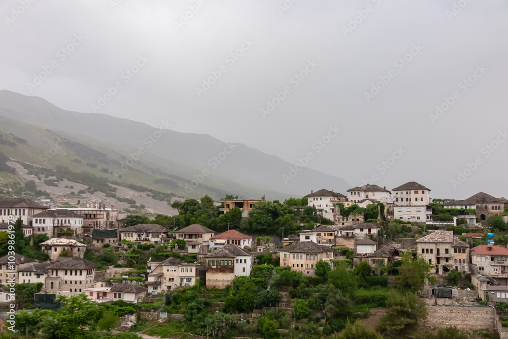 Fototapeta premium Panoramic view of the city of Gjirokaster, Albania. Nestled in valley between mountains, with a dense cluster of whitewashed houses with traditional layered stone tile roofs. Historical charm. Tourism
