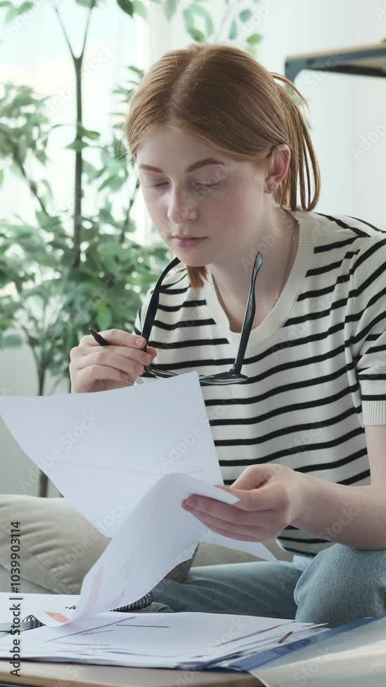 Thoughtful redhead Caucasian girl student doing homework with papers at home. Concept of a remote learning or working. Vertical video. Thoughtful redhead Caucasian girl student doing homework with papers at home. Concept of a remote learning or working. Vertical video.