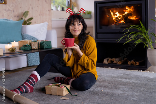 Cozy woman enjoying hot drink by fireplace, wrapping Christmas gifts at home photo