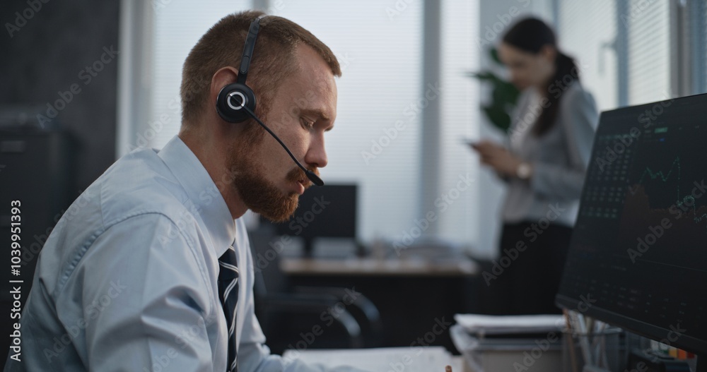 Professional stock exchange trader works in modern office. Financial specialist in headset works on computer, talks to clients, monitors share markets, takes notes. Female colleague in the background. Professional stock exchange trader works in modern office. Financial specialist in headset works on computer, talks to clients, monitors share markets, takes notes. Female colleague in the background.