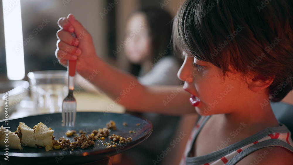 Fototapeta premium Young boy eating at the dining table, holding a fork and focusing on the food on his plate during meal time in a relaxed home setting