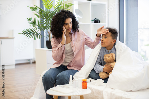 African-American mother checks sick son's temperature while speaking to doctor on phone. Young boy lies on bed wrapped in blanket holding teddy bear, showing concern and care from parent.