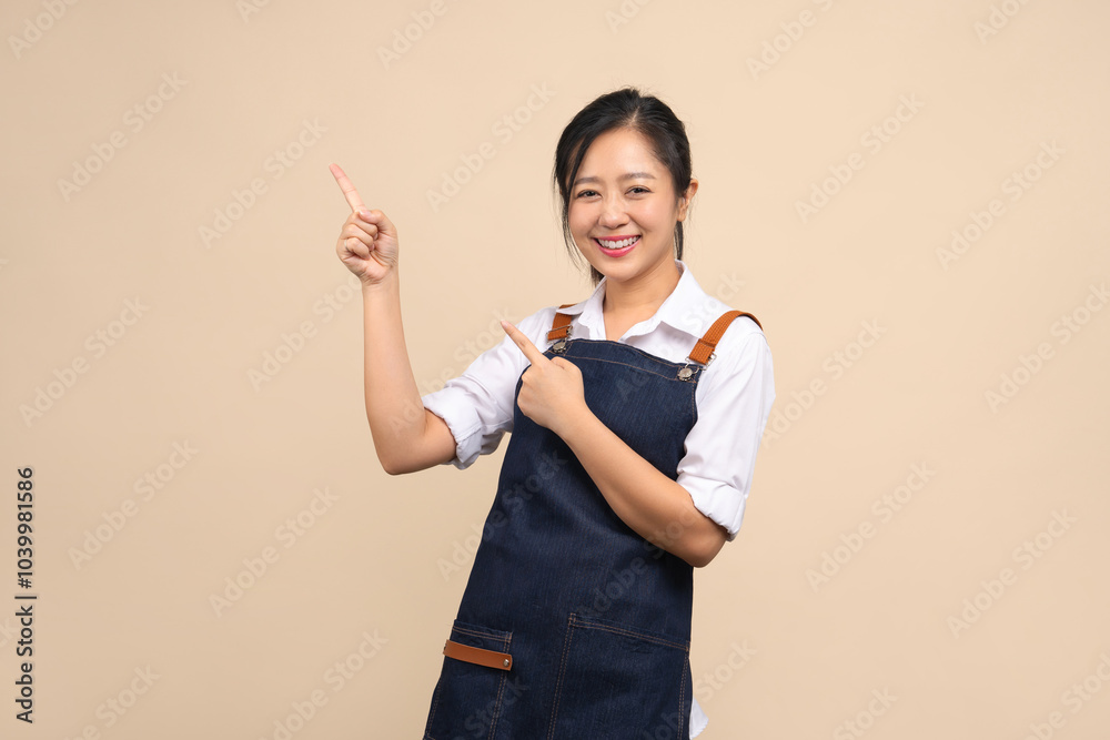 Cute Asian woman in apron pointing her finger to show space for your ad on beige background.