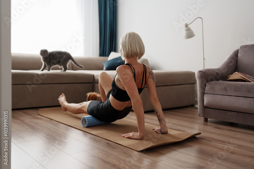 A woman performs foam rolling on a yoga mat in her living room, while her cat looks on. The scene combines fitness with the comfort of home life.