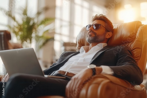 A businessman sits comfortably in an upscale leather chair, wearing sunglasses and looking relaxed. He has a laptop open on his lap and enjoys the serene atmosphere of a stylish office lounge illumina photo