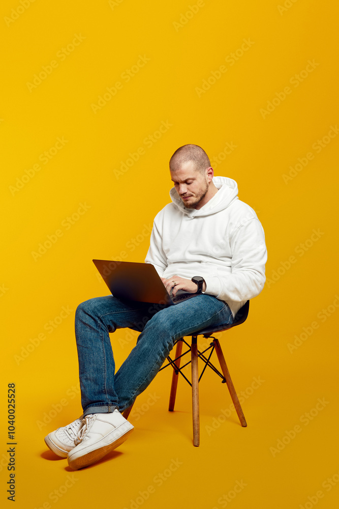 Vertical photo of full body young man sitting on chair and using modern notebook, typing on laptop, isolated on yellow background Vertical photo of full body young man sitting on chair and using modern notebook, typing on laptop, isolated on yellow background