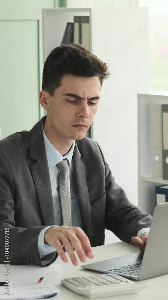 Serious young adult Caucasian employee working with laptop and calculator in the office, planning strategy or analyzing finances. Vertical video. Serious young adult Caucasian employee working with laptop and calculator in the office, planning strategy or analyzing finances. Vertical video.