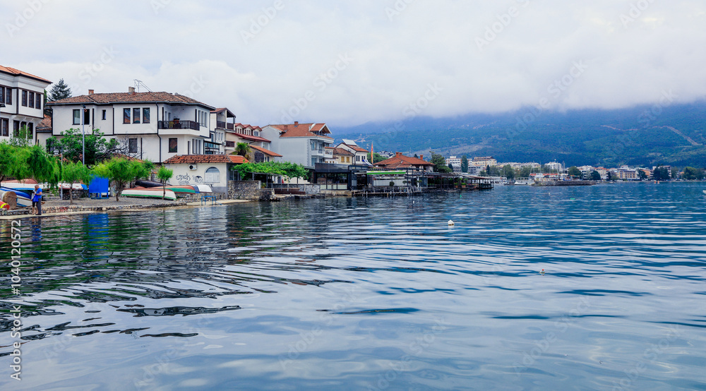 A serene view of Ohrid\'s lakefront homes reflecting on the water during a cloudy day in Macedonia A serene view of Ohrid\'s lakefront homes reflecting on the water during a cloudy day in Macedonia