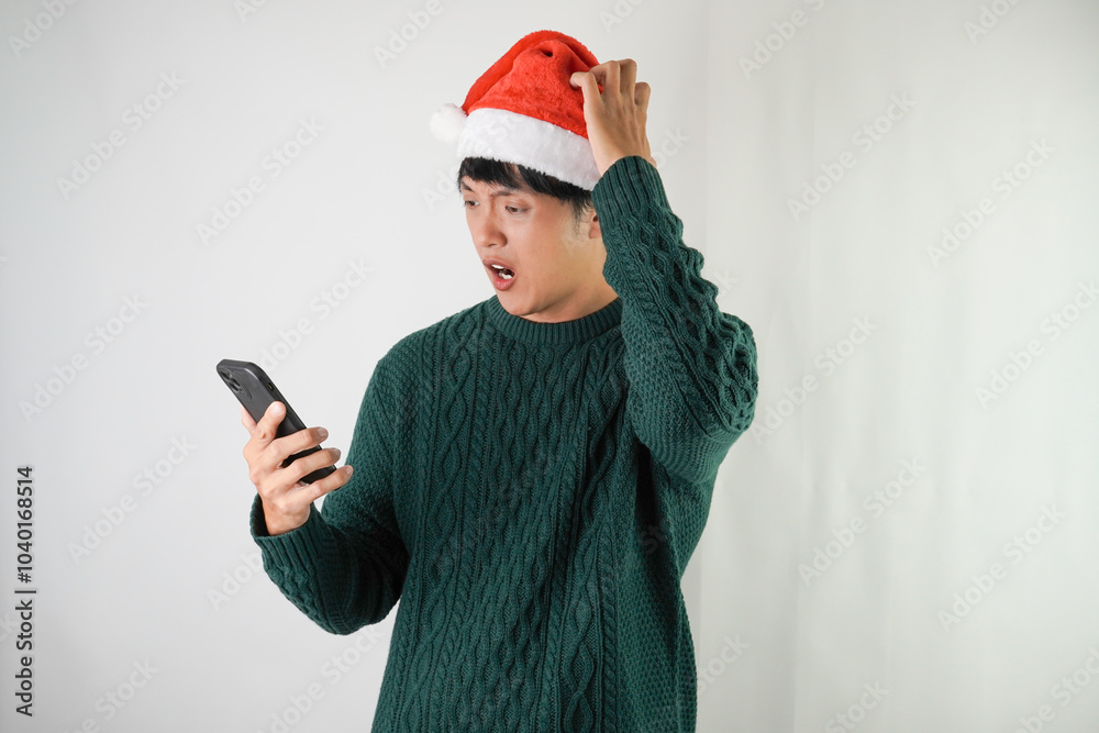 Angry young asian man wearing santa clause hat and green long sleeved sweater is using smartphone, isolated over white background. Concept for Christmas Holiday and New Year Party Angry young asian man wearing santa clause hat and green long sleeved sweater is using smartphone, isolated over white background. Concept for Christmas Holiday and New Year Party