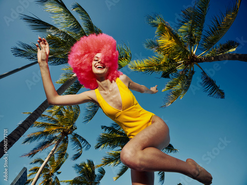 Woman, yellow swimsuit, pink wig, jumping, palm trees a woman in a vibrant yellow swimsuit and playful pink wig is captured midjump with a backdrop of lush palm trees and clear blue skies photo