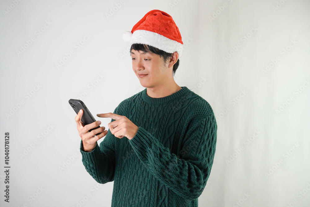Excited young asian man wearing santa clause hat and green long sleeved sweater is using smartphone, isolated over white background. Concept for Christmas Holiday and New Year Party Excited young asian man wearing santa clause hat and green long sleeved sweater is using smartphone, isolated over white background. Concept for Christmas Holiday and New Year Party