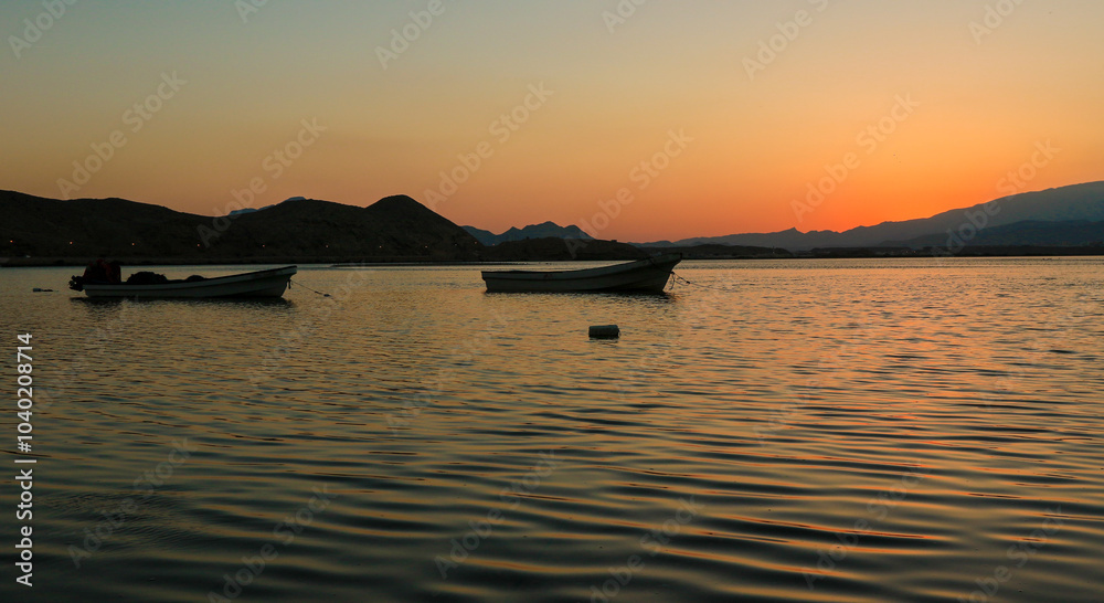 Sunset over the tranquil waters of Sur Oman featuring traditional boats against a backdrop of mountains Sunset over the tranquil waters of Sur Oman featuring traditional boats against a backdrop of mountains