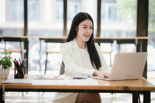 Beautiful Asian female accountant working with smartphone and laptop at her desk analyzing business reports and documents. Sending messages