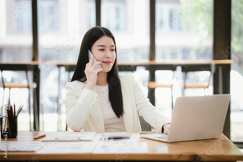 Beautiful Asian female accountant working with smartphone and laptop at her desk analyzing business reports and documents. Sending messages