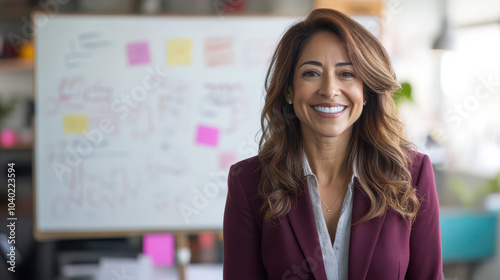 Energetic Hispanic female marketing manager in burgundy blazer presenting ideas on whiteboard. Three-quarter portrait in a bright creative workspace. Professional and engaging image.