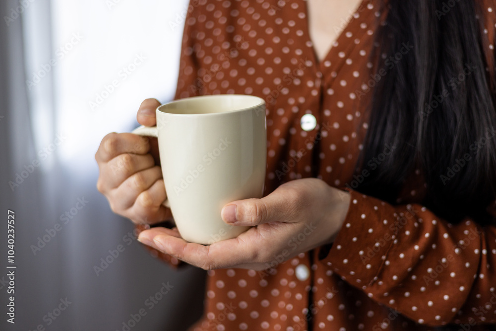 Close-up of asian woman holding mug, displaying comfort, warmth, and relaxation. Hands cradling neutral-colored cup, wearing red polka dot blouse, suggesting cozy, calm atmosphere. Close-up of asian woman holding mug, displaying comfort, warmth, and relaxation. Hands cradling neutral-colored cup, wearing red polka dot blouse, suggesting cozy, calm atmosphere.