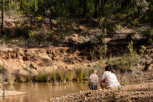 Teen girl with her young niece exploring australian bush squatting down by waterhole in bushland
