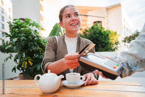 Smiling young woman sitting at a bar terrace, paying for her tea using a credit card and contactless payment terminal, highlighting convenience and modern technology in outdoor dining settings photo