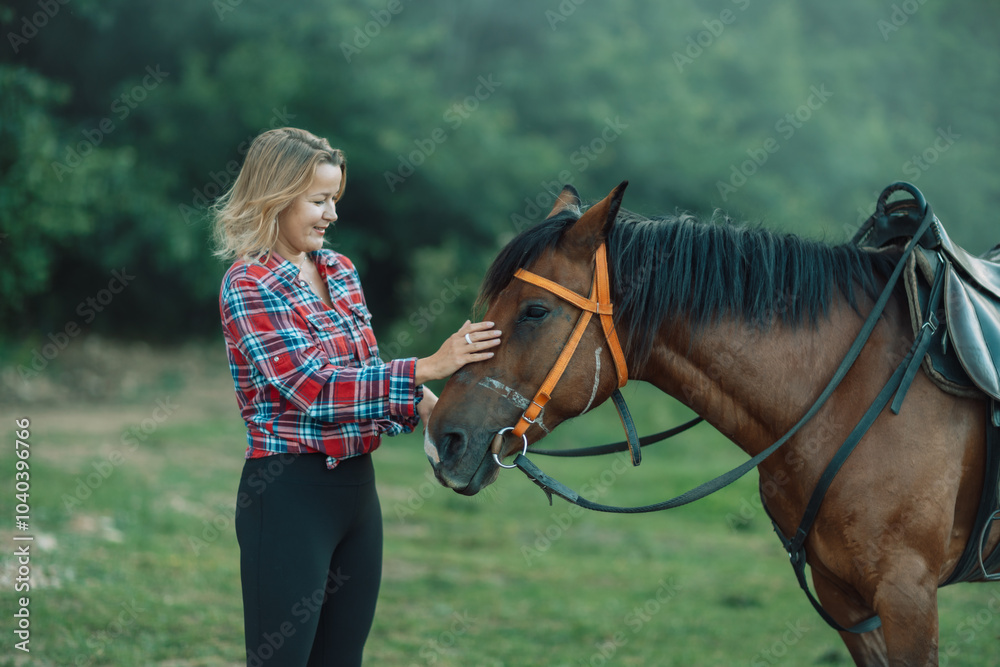 Obraz premium Happy blonde with horse in forest. Woman and a horse walking through the field during the day. Dressed in a plaid shirt and black leggings.