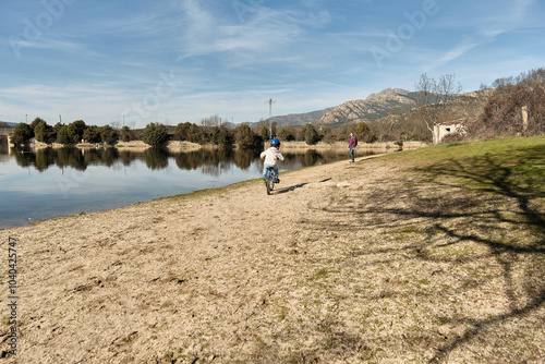 A boy rides his bicycle towards his mother along a path by a lake.