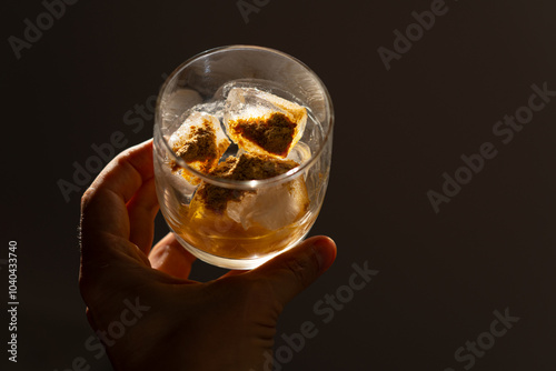 A glass filled with ice cubes and powdered Amanita muscaria mushroom on its surface, held in the hand, illuminated by natural sunlight. Preparing a homemade fly agaric elixir photo