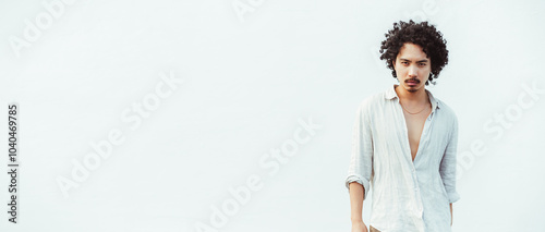 Young asian man with curly hair wearing a light, unbuttoned linen shirt stands confidently against a clean, white background. Ample copy space area to the left photo