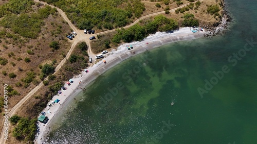 A stunning aerial view of a coastal landscape featuring a serene bay, vast green fields, and distant mountains under a clear blue sky. The image captures a peaceful seaside area with a few ships