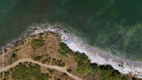 A stunning aerial view of a coastal landscape featuring a serene bay, vast green fields, and distant mountains under a clear blue sky. The image captures a peaceful seaside area with a few ships