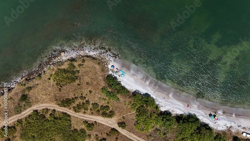 A stunning aerial view of a coastal landscape featuring a serene bay, vast green fields, and distant mountains under a clear blue sky. The image captures a peaceful seaside area with a few ships