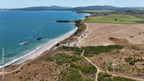 A stunning aerial view of a coastal landscape featuring a serene bay, vast green fields, and distant mountains under a clear blue sky. The image captures a peaceful seaside area with a few ships