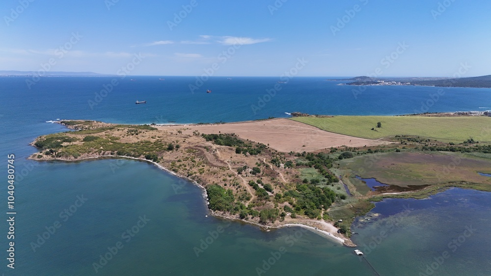 Obraz premium A stunning aerial view of a coastal landscape featuring a serene bay, vast green fields, and distant mountains under a clear blue sky. The image captures a peaceful seaside area with a few ships
