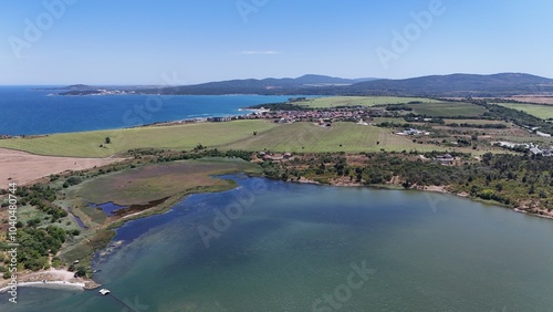 A stunning aerial view of a coastal landscape featuring a serene bay, vast green fields, and distant mountains under a clear blue sky. The image captures a peaceful seaside area with a few ships