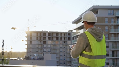 Man constructive engineer with white hard hat and safety vest is using a tablet computer while inspecting a construction site at sunset