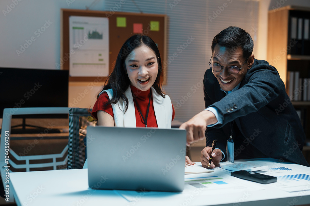 Two coworkers are looking happy while working late on a laptop in their office at night Two coworkers are looking happy while working late on a laptop in their office at night