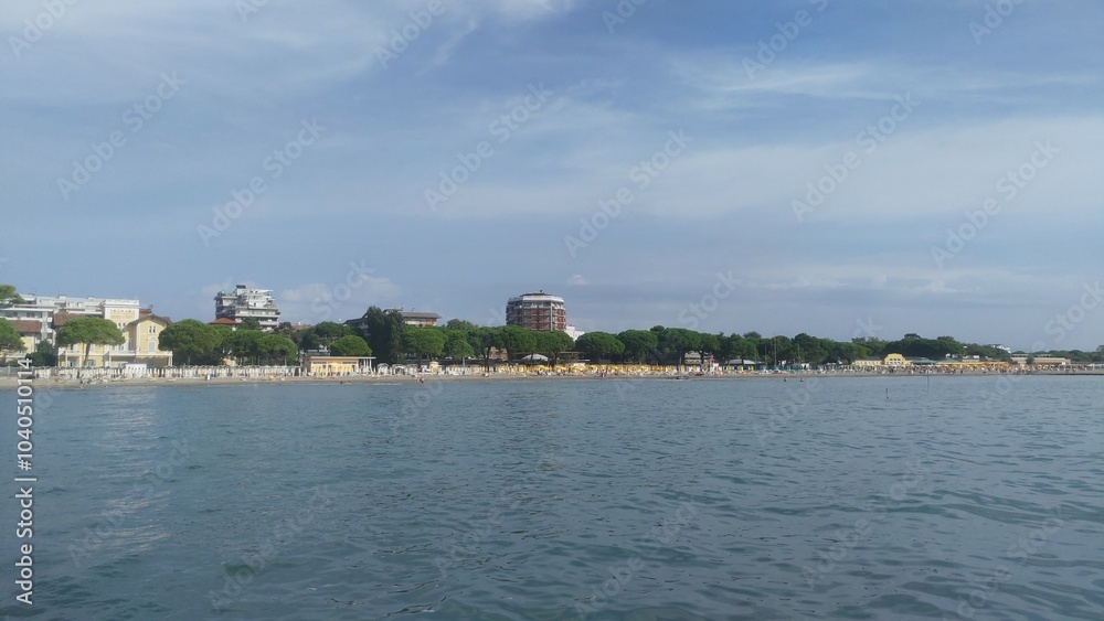 On the sandy beach under the pine trees there are plastic sunbeds and umbrellas. People rest on them and swim in the sea. Behind the beach are buildings. Sunny autumn weather and blue sky with clouds On the sandy beach under the pine trees there are plastic sunbeds and umbrellas. People rest on them and swim in the sea. Behind the beach are buildings. Sunny autumn weather and blue sky with clouds