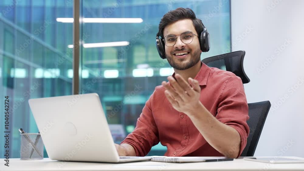 Smiling IT specialist in wireless headphones asks to join him, beckons with inviting hand gesture while sitting at laptop at workplace in office. Friendly developer or coder invites to join the team Smiling IT specialist in wireless headphones asks to join him, beckons with inviting hand gesture while sitting at laptop at workplace in office. Friendly developer or coder invites to join the team