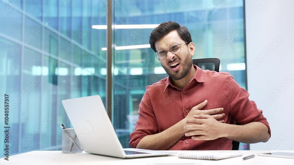 Handsome businessman has a heart attack while working on a laptop while sitting at workplace in business office. A sick worker suffers from tension in the chest and holds the sore spot with his hands Handsome businessman has a heart attack while working on a laptop while sitting at workplace in business office. A sick worker suffers from tension in the chest and holds the sore spot with his hands