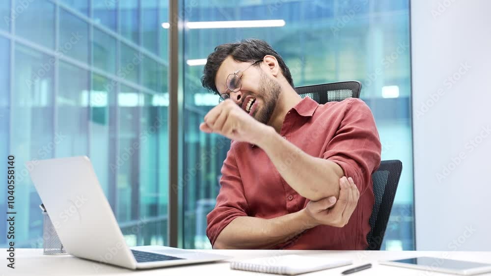 Businessman suffering from elbow pain while working on a laptop sitting at desk in a business office. Upset male is massaging a joint, performs hand exercises. He has a joint injury or ligament sprain Businessman suffering from elbow pain while working on a laptop sitting at desk in a business office. Upset male is massaging a joint, performs hand exercises. He has a joint injury or ligament sprain