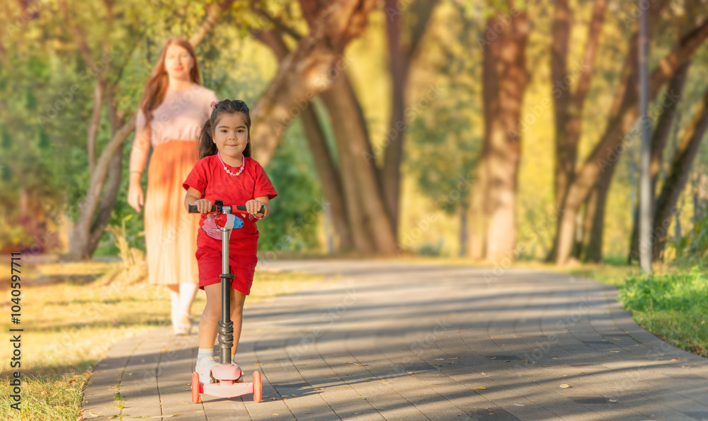 A little girl on a scooter in the park with her mother. The sun is shining brightly. A little girl on a scooter in the park with her mother. The sun is shining brightly.