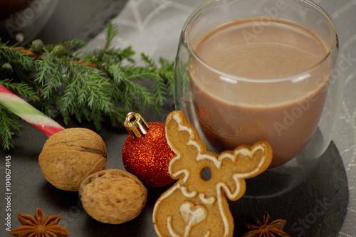 
New Year's composition. Horizontal photo. A clear glass cup with hot cocoa poured into it. Near the table there is a pine branch, a gingerbread deer, candy canes, nuts, fir decorations, and anise.