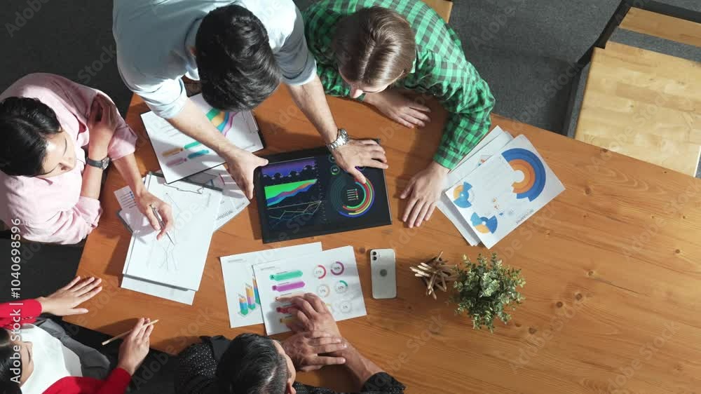Top view of business man using tablet display increasing graph while diverse marketing people sharing idea or planing strategy at meeting table with stock market statistic scatter around. Convocation. Top view of business man using tablet display increasing graph while diverse marketing people sharing idea or planing strategy at meeting table with stock market statistic scatter around. Convocation.